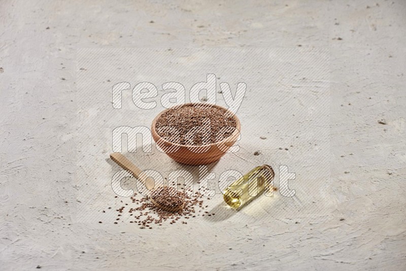 A wooden bowl and spoon full of flaxseeds with a bottle of flaxseeds oil on a textured white flooring