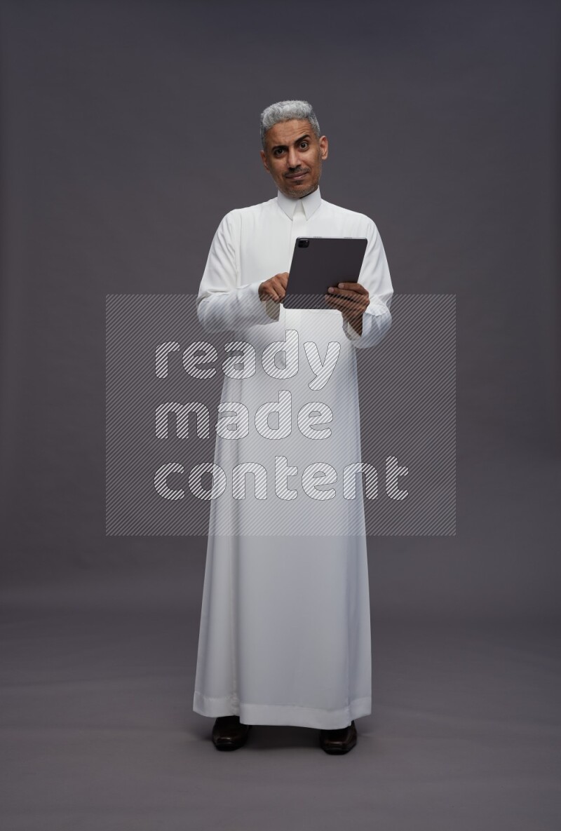 Saudi man wearing thob standing working on tablet on gray background