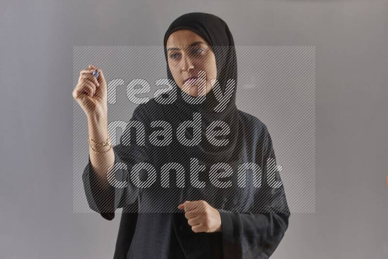 Woman in black abaya and hijab holding a marker pen to write on a transparent board with different reactions and expressions on grey background