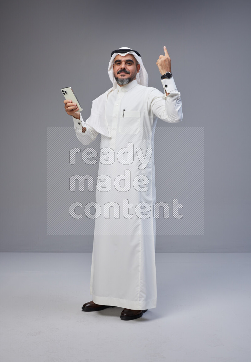Saudi man Wearing Thob and white Shomag standing texting on phone on Gray background