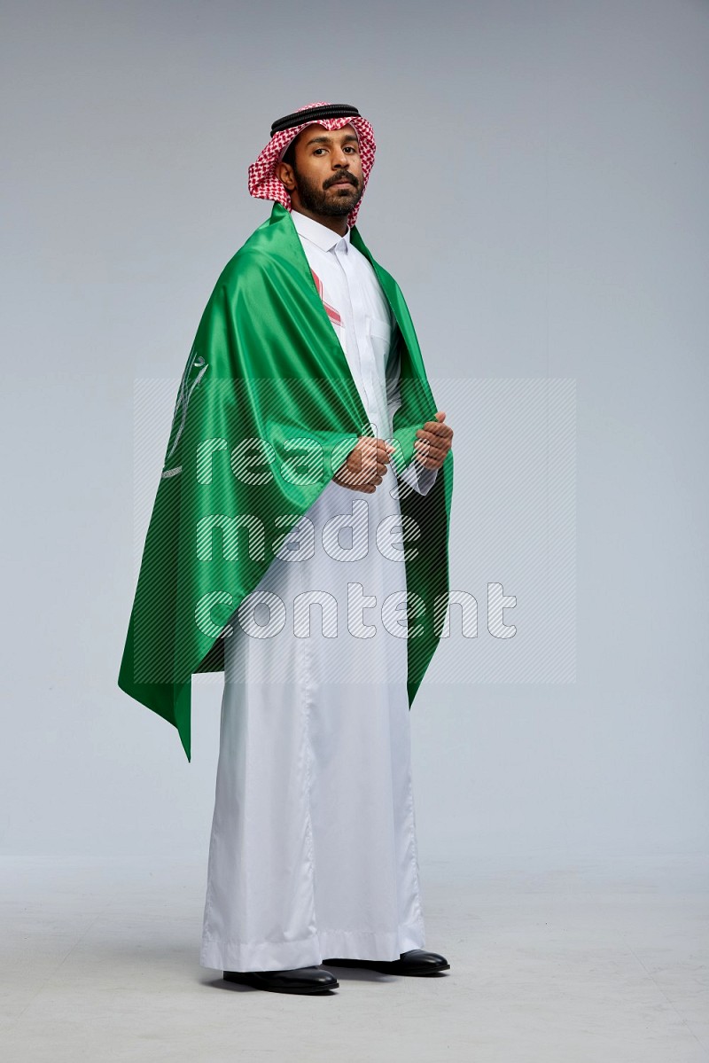 Saudi man Wearing Thob and shomag standing holding Saudi flag on Gray background