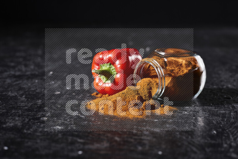 A glass jar full of ground paprika powder flipped with some spilling powder on black background