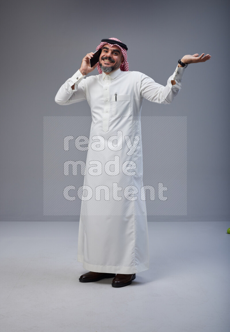 Saudi man Wearing Thob and red Shomag standing talking on phone on Gray background