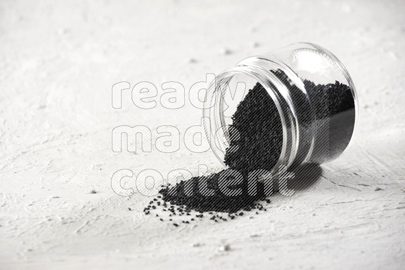 A glass jar full of black seeds and the jar flipped and seeds spread on a textured white flooring