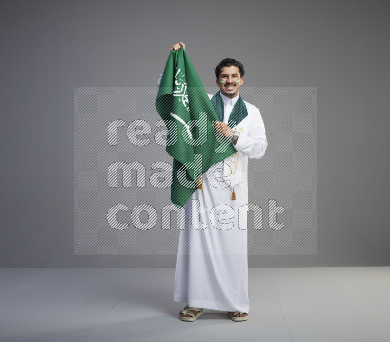 A Saudi man standing wearing thob with face painting wrapping Saudi flag scarf and holding big Saudi flag on gray background