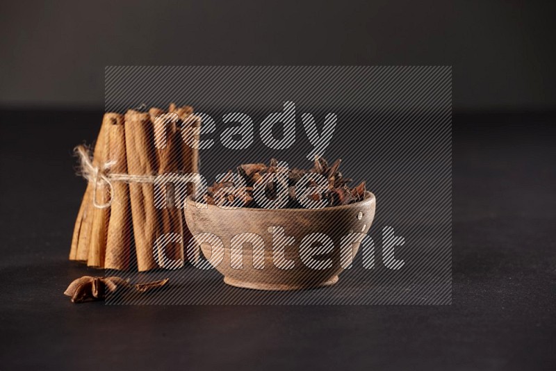A stacked and bounded cinnamon sticks and a wooden bowl full of star anise on a black background