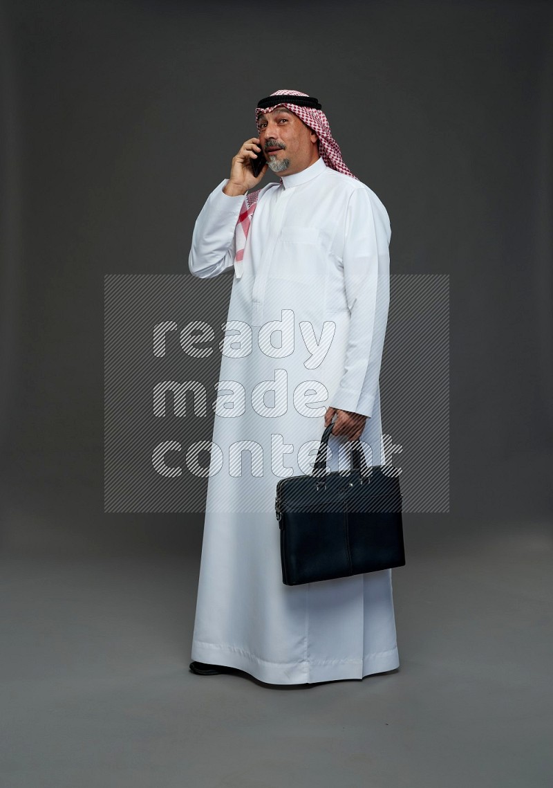 Saudi man with shomag Standing holding bag talking on phone on gray background