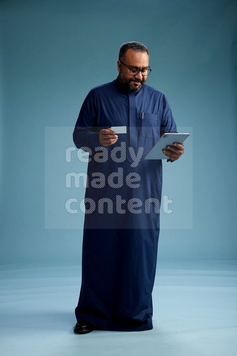Saudi Man without shimag Standing holding ATM card while working on tablet on blue background