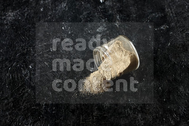 A flipped glass jar full of cardamom powder and powder spilled out of it on textured black flooring