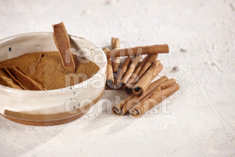 Ceramic bowl full of cinnamon powder with cinnamon sticks on the side on white background