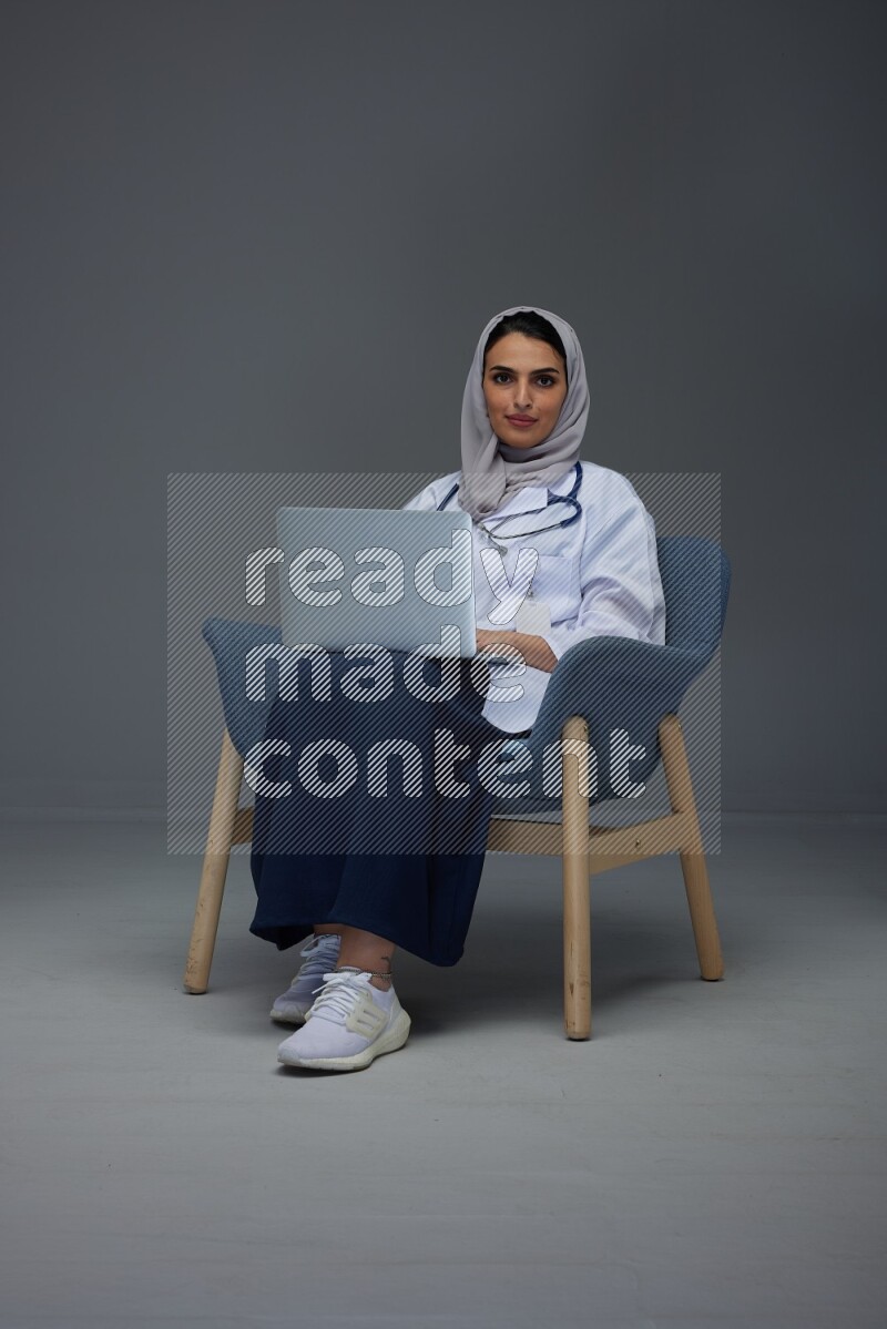 A doctor wearing a light gray head scarf sitting on a dark grey chair and using a laptop eye level on a grey background