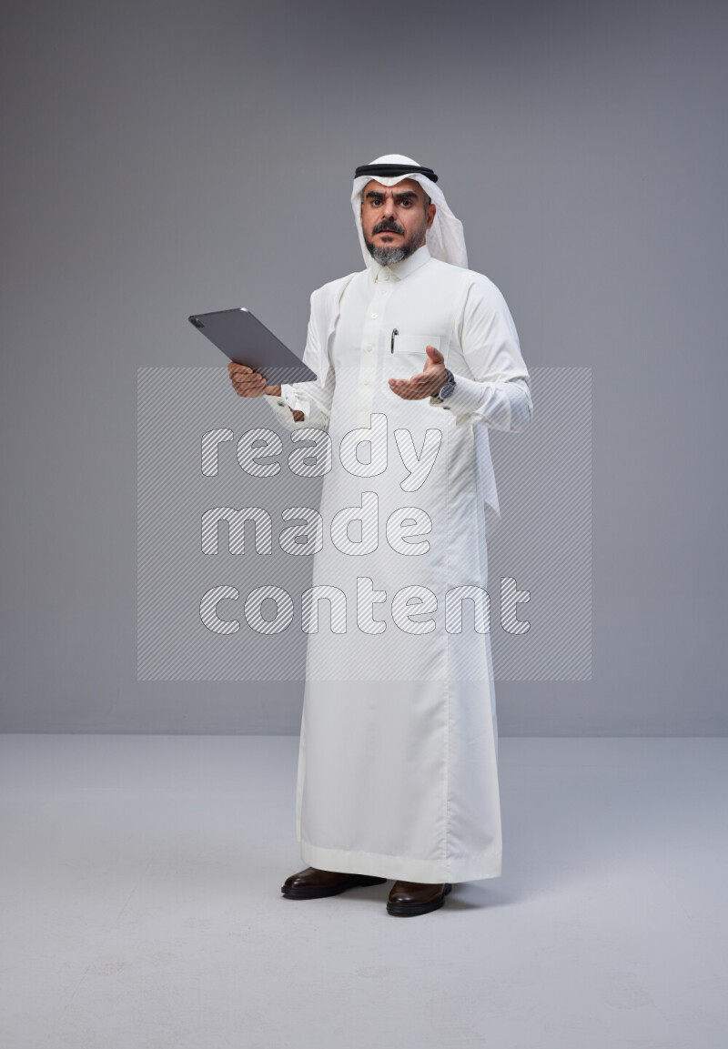 Saudi man Wearing Thob and white Shomag standing working on tablet on Gray background