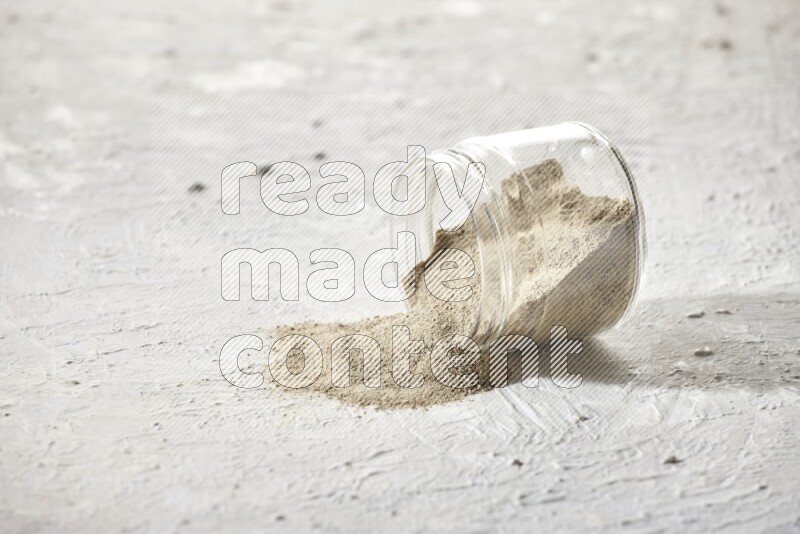 A flipped glass jar full of white pepper powder with spilled powder on textured white flooring