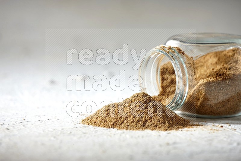 A flipped glass spice jar full of cumin powder and powder spilled out on textured white flooring