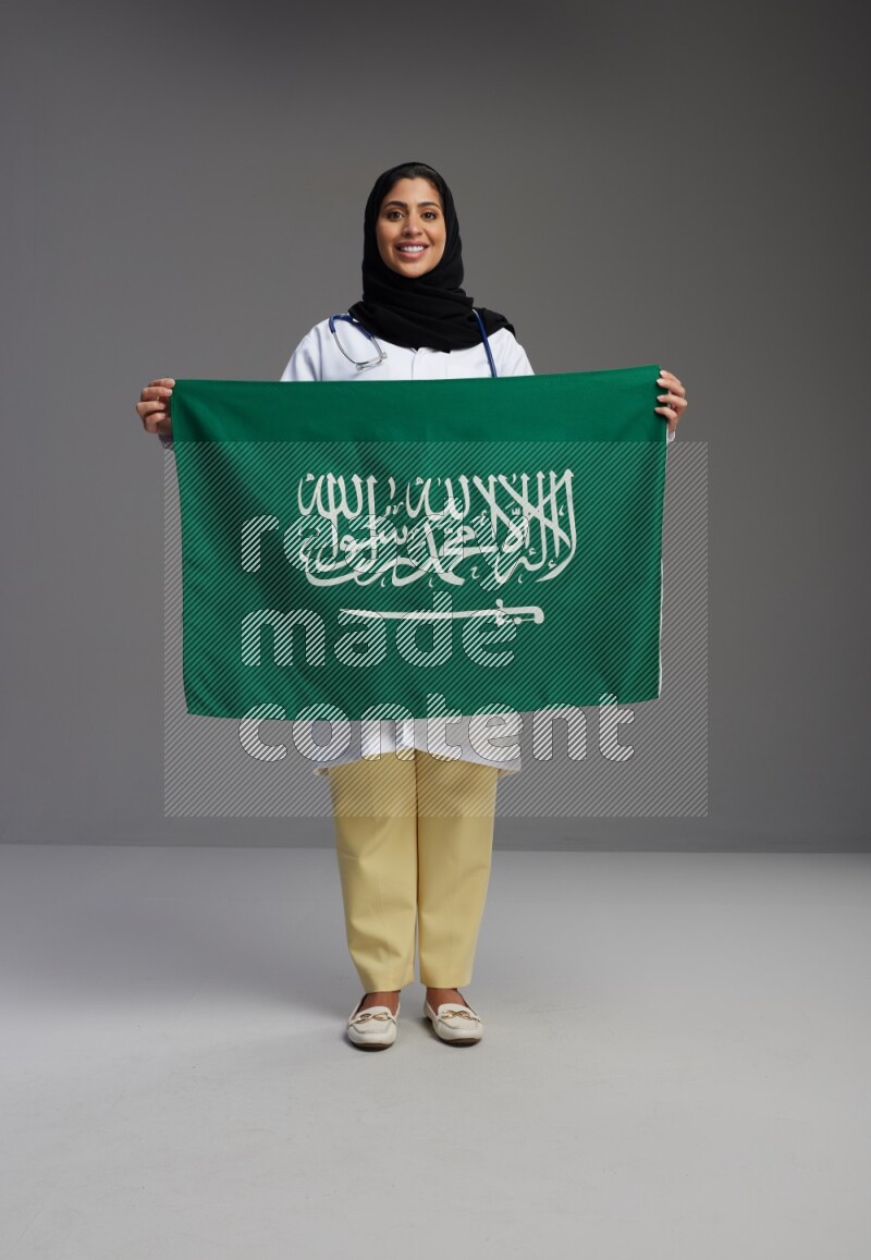 Saudi woman wearing lab coat with stethoscope standing holding Saudi flag on Gray background