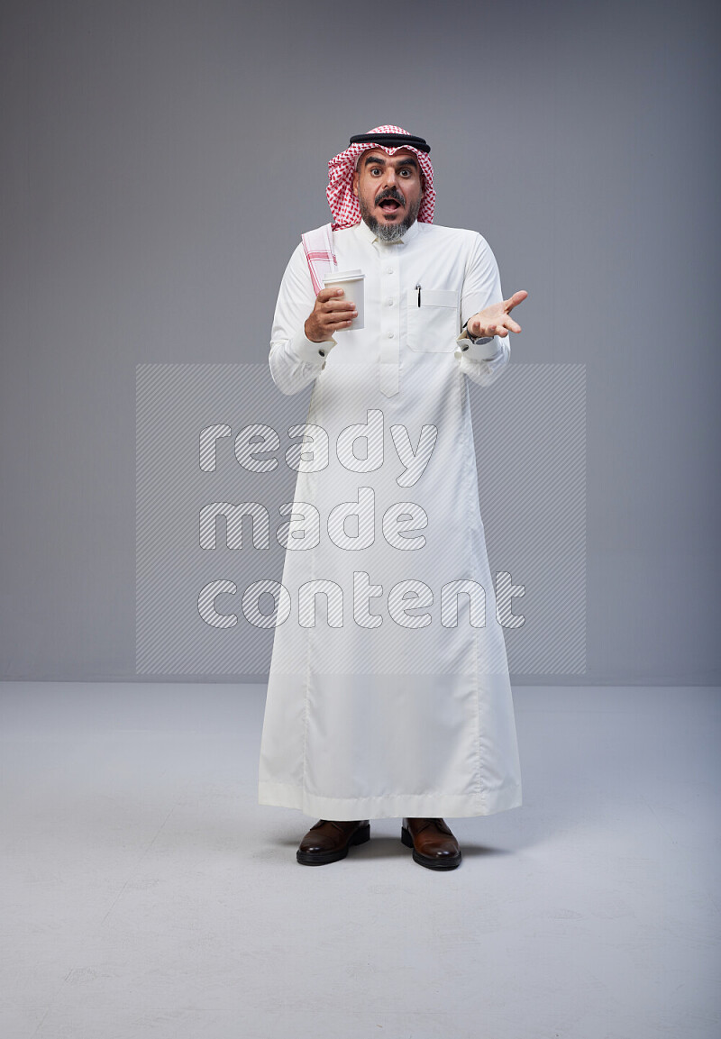 Saudi man Wearing Thob and red Shomag standing holding paper cup on Gray background