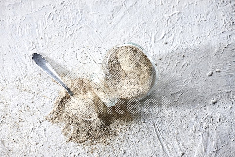 A flipped herbal glass jar and metal spoon full of white pepper powder with spilled powder on textured white flooring