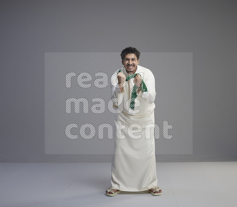 A Saudi man standing wearing thob holding Saudi flag scarf on gray background
