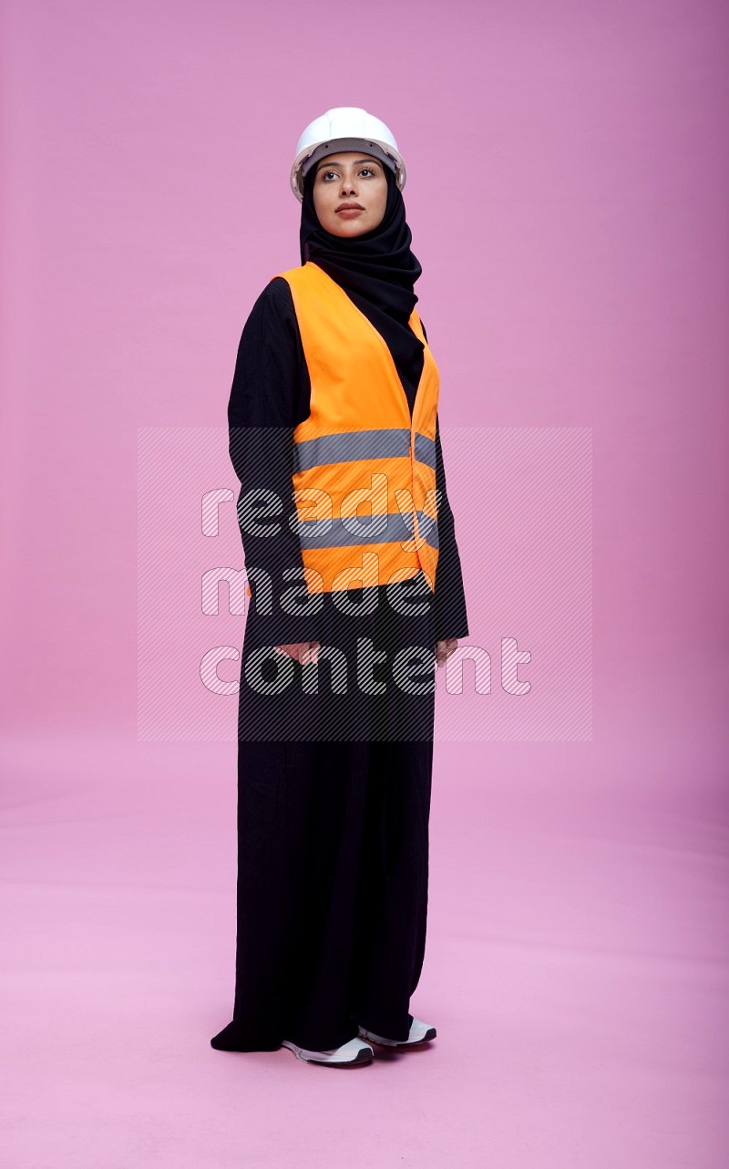 Saudi woman wearing Abaya with engineer vest and helmet standing interacting with the camera on pink background