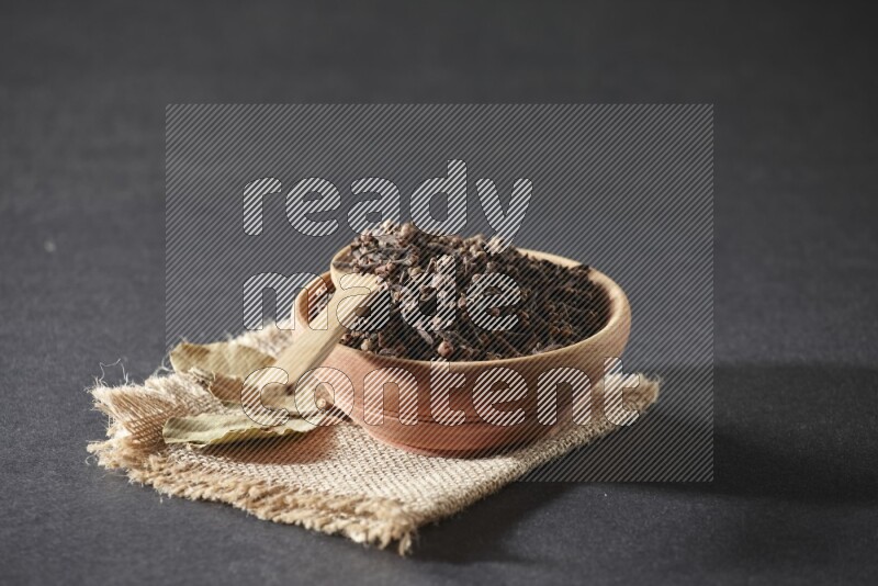 A wooden bowl, a wooden spoon full of cloves, and bay leaves (laurel) on a piece of burlap on a black flooring