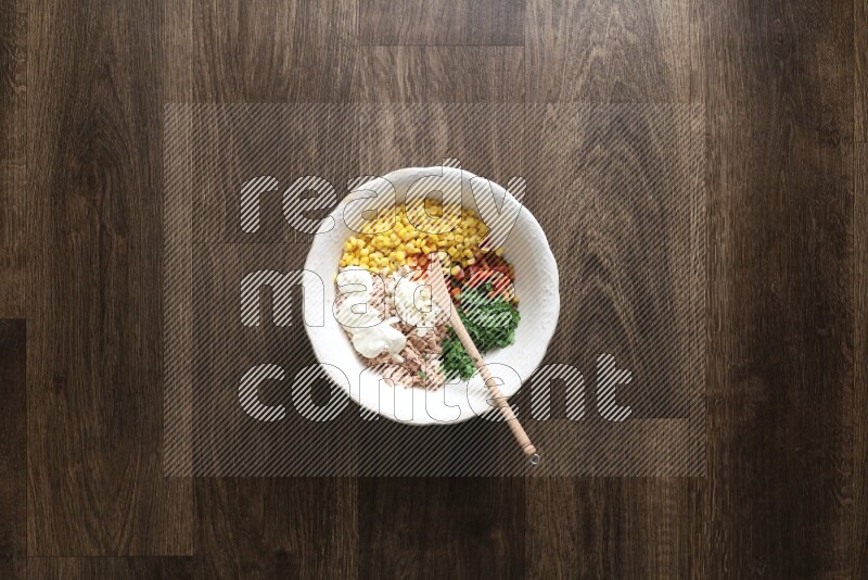 A white bowl full of tuna, colored bell pepper, sour cream, corn, parsley, black pepper powder and sauce, with wooden spoon on wooden background