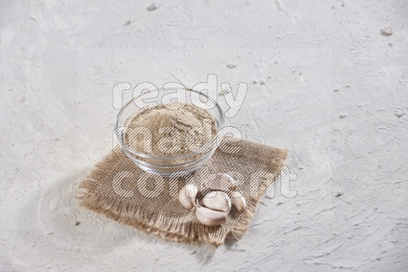 A glass bowl full of garlic powder placed on burlap fabric with garlic cloves on a textured white flooring