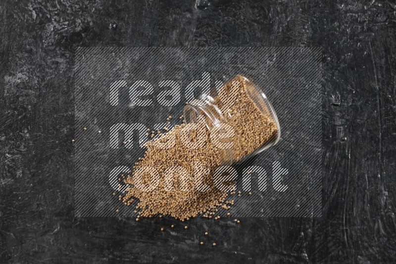 A glass jar full of mustard seeds and jar is flipped and seeds spread out on a textured black flooring