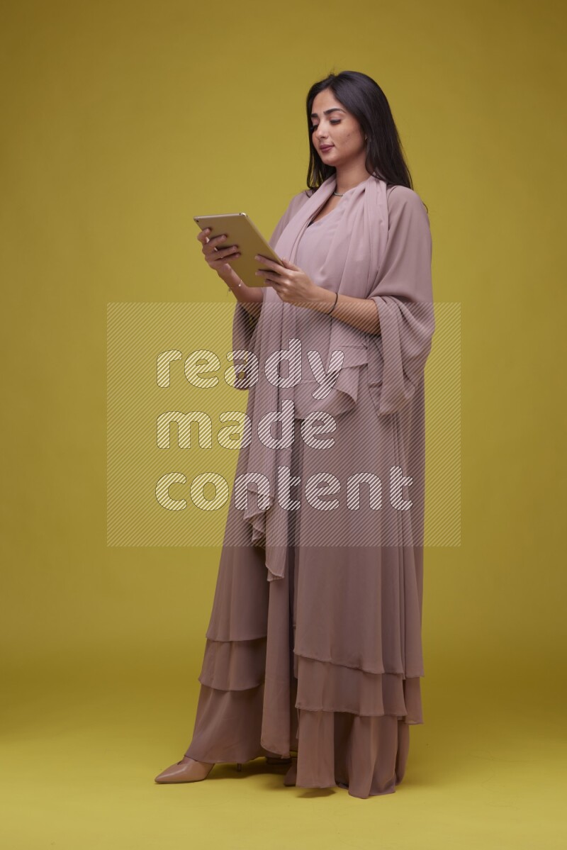 A Saudi woman Holding an iPad  on a Yellow Background wearing Brown Abaya