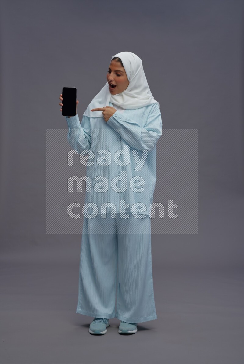 Saudi woman wearing hijab clothes standing showing phone to camera on gray background