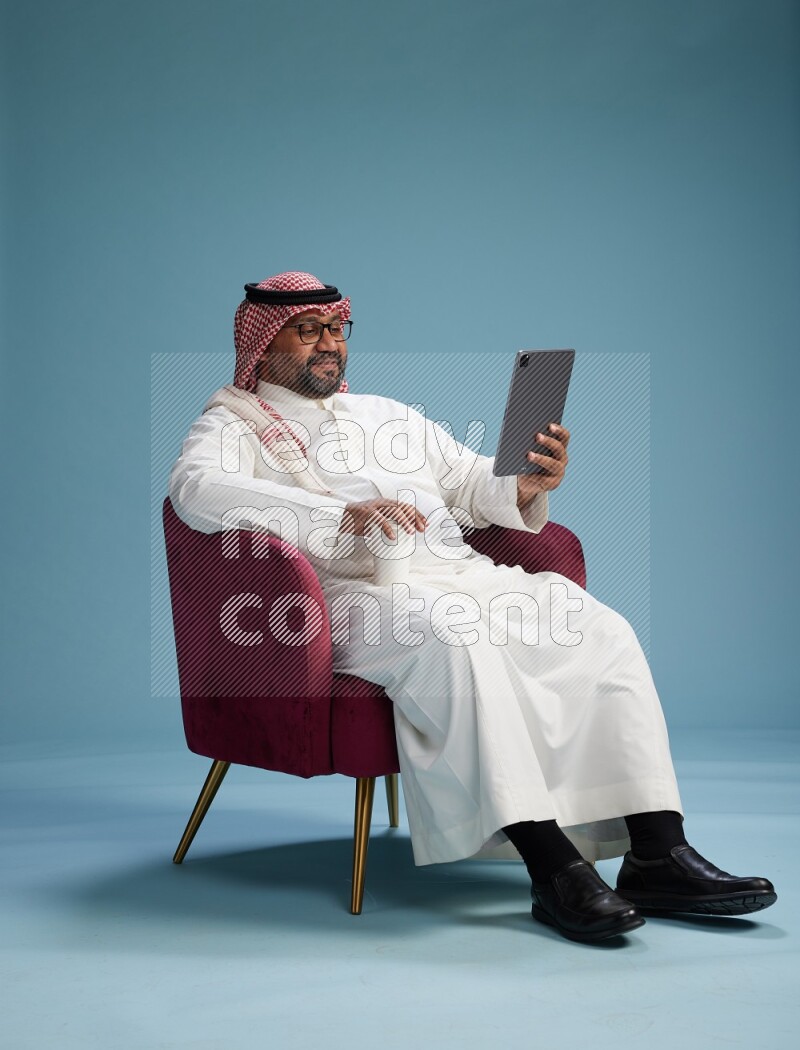 Saudi Man with shimag sitting on chair working on tablet on blue background