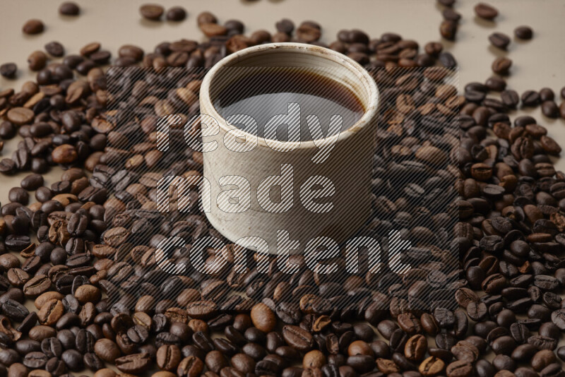 A beige pottery cup of coffee surrounded by roasted coffee beans on beige background