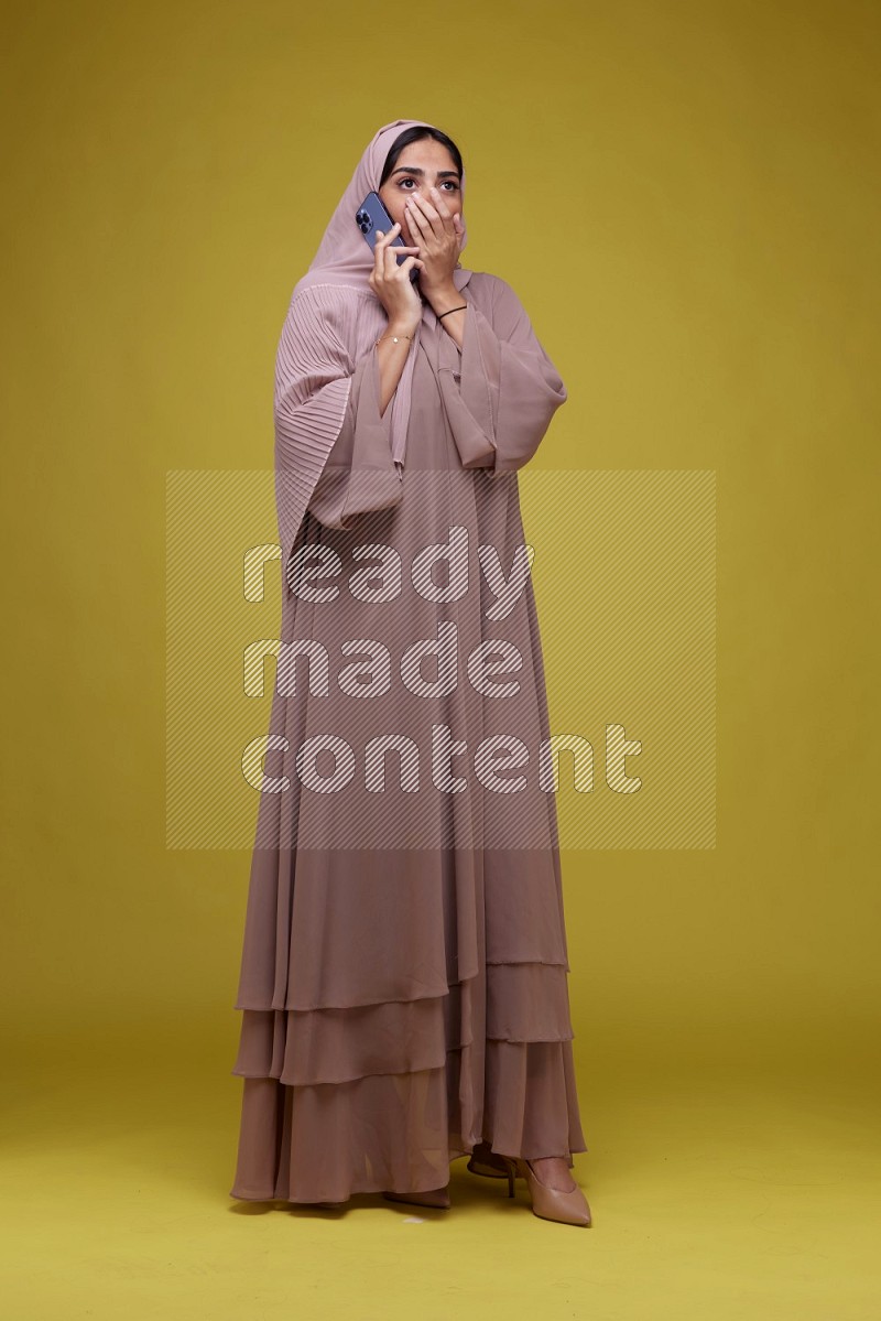 A woman having a call on a Yellow Background wearing Brown Abaya with Hijab