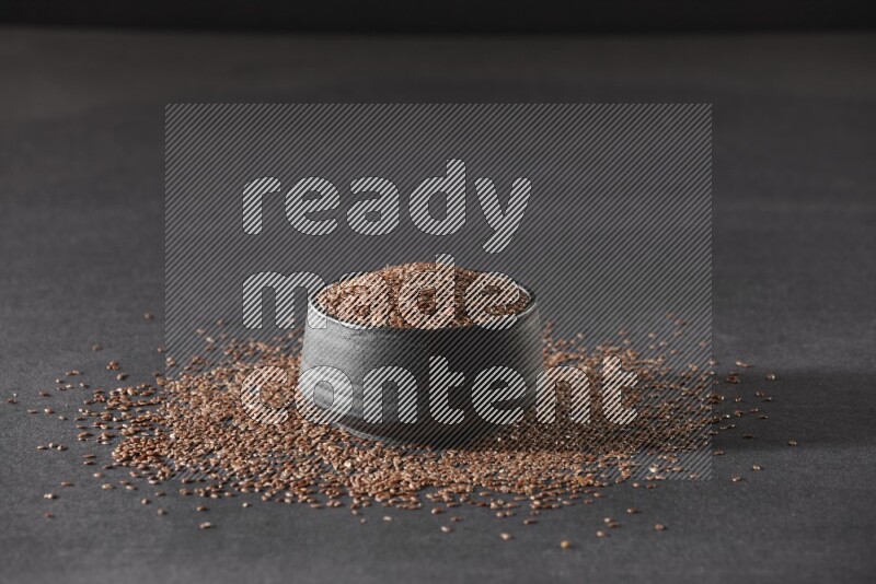 A black pottery bowl full of flaxseeds surrounded by the seeds on a black flooring