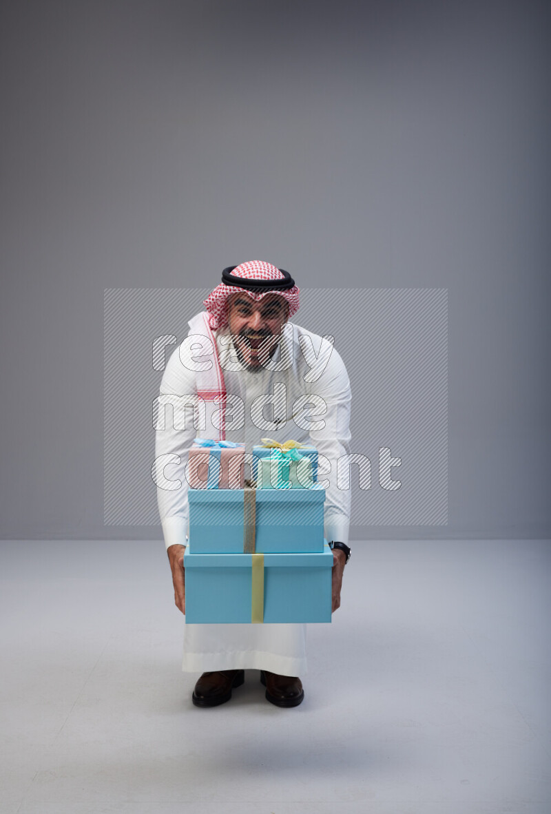 Saudi man Wearing Thob and red Shomag standing holding gift box on Gray background