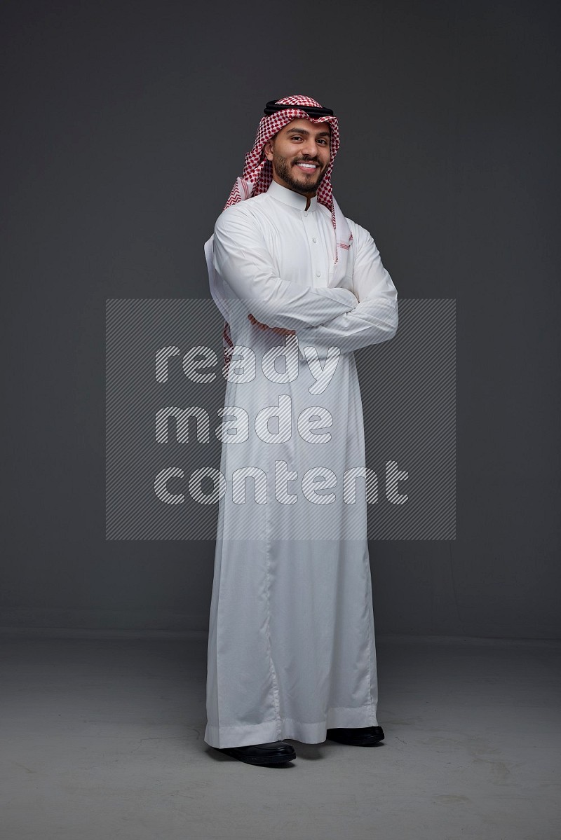 A Saudi man wearing Thobe and Shmagh standing and using his phone eye level on a gray background