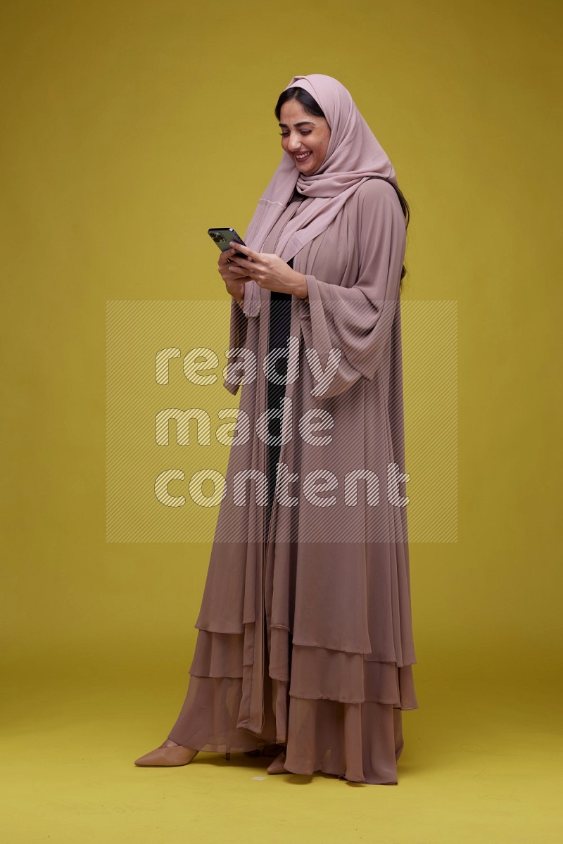 A woman Texting on her phone on a Yellow Background wearing Brown Abaya with Hijab