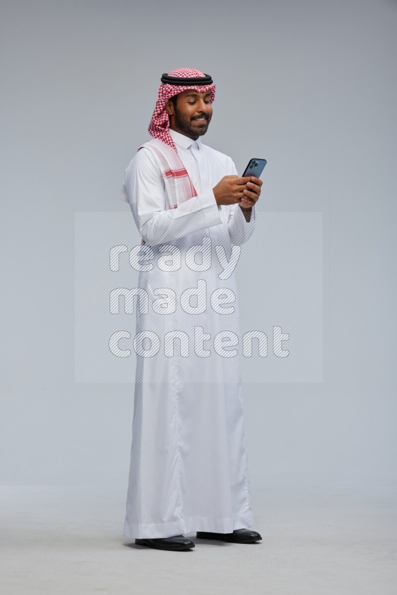 Saudi man Wearing Thob and shomag standing texting on phone on Gray background