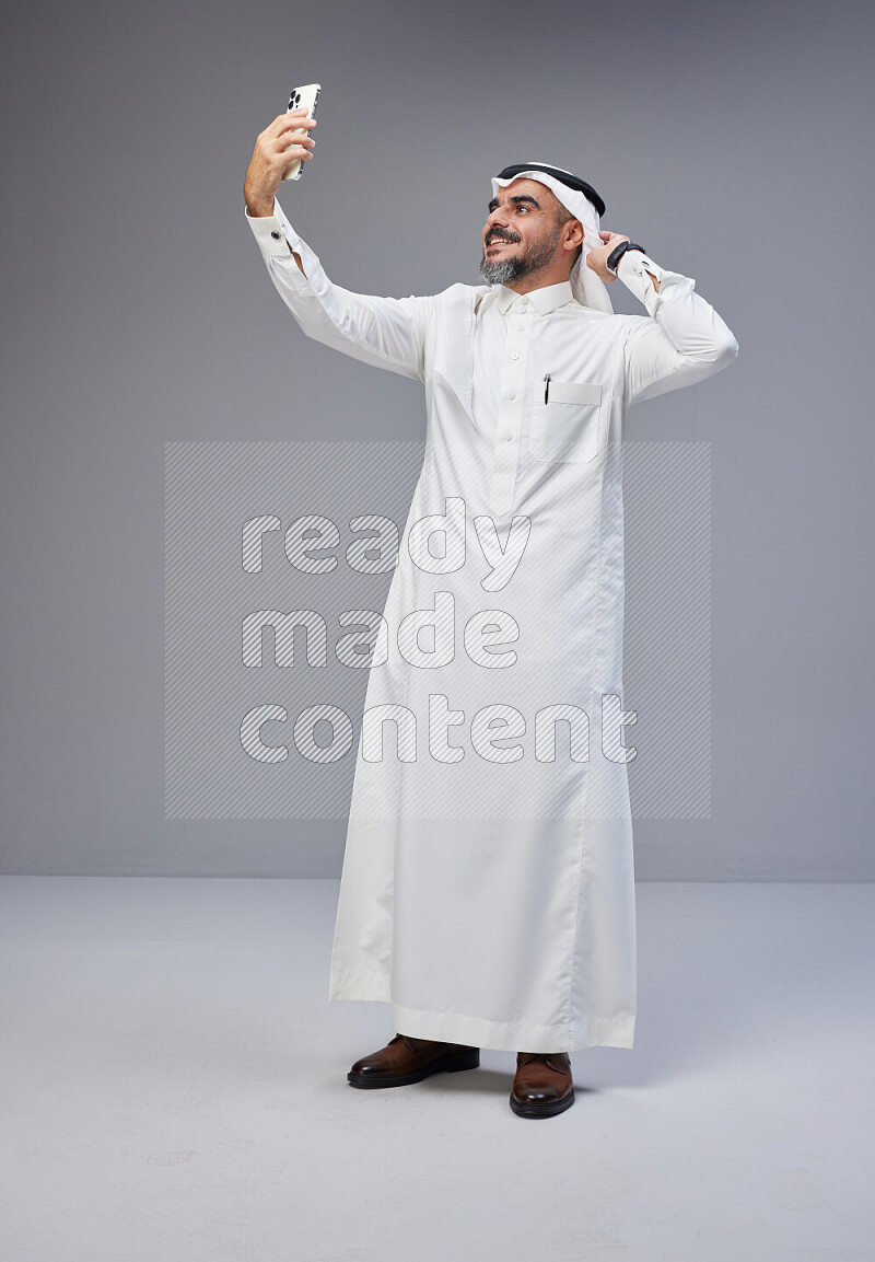 Saudi man Wearing Thob and white Shomag standing taking selfie on Gray background