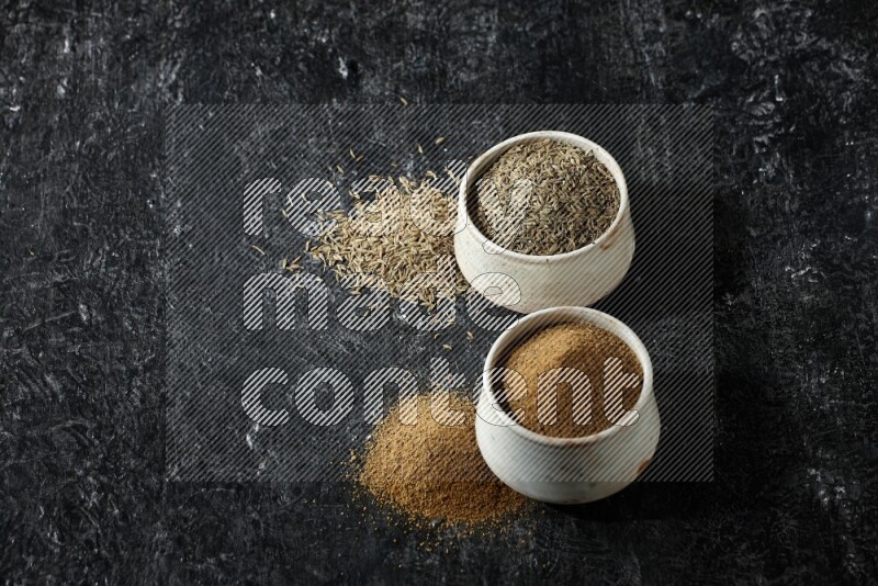 2 beige bowls, one full of cumin powder and the other full of seeds on a textured black flooring