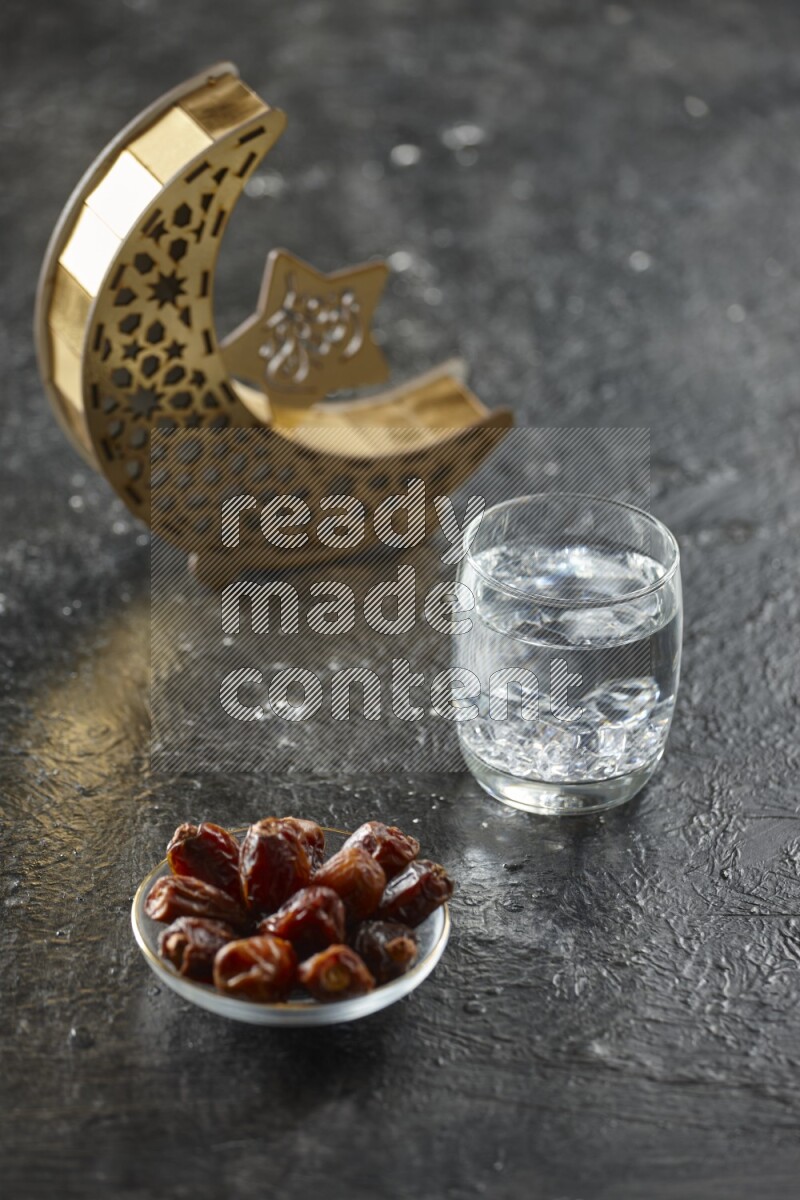 A wooden golden crescent lantern with different drinks, dates, nuts, prayer beads and quran on textured black background