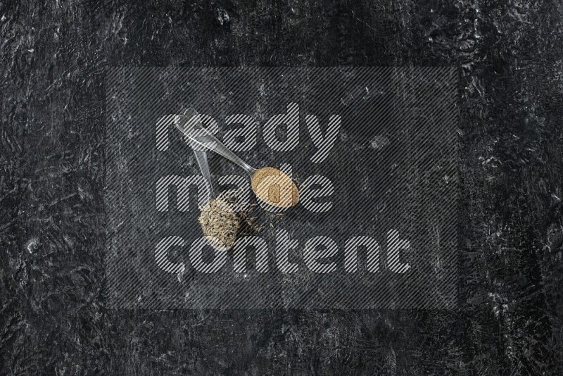 ملعقتان معدنيتان مملوءتان بحبوب الكمون وبودرة الكمون على أرضية سوداء