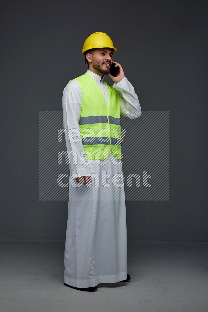 A Saudi man wearing Thobe with a yellow safety vest and white helmet standing and talking in the phone eye level on a gray background