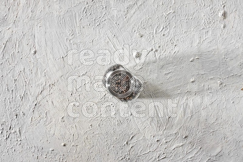 A glass spice jar full of black pepper on textured white flooring