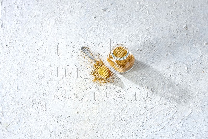 وعاء زجاجي للتوابل وملعقة معدنية ممتلئان ببودرة الكركم على خلفية بيضاء