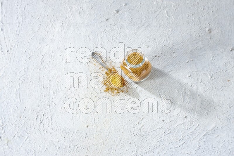 A glass spice jar and metal spoon full of turmeric powder on textured white flooring