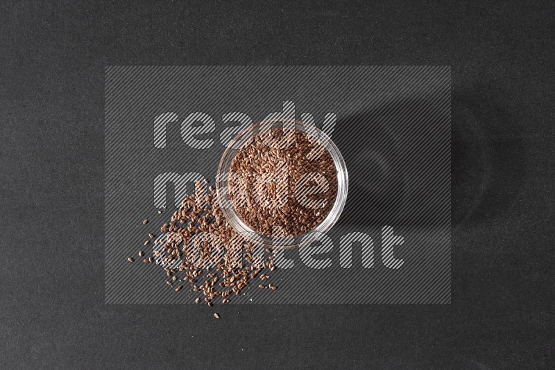 A glass bowl full of flaxseeds surrounded by the seeds on a black flooring