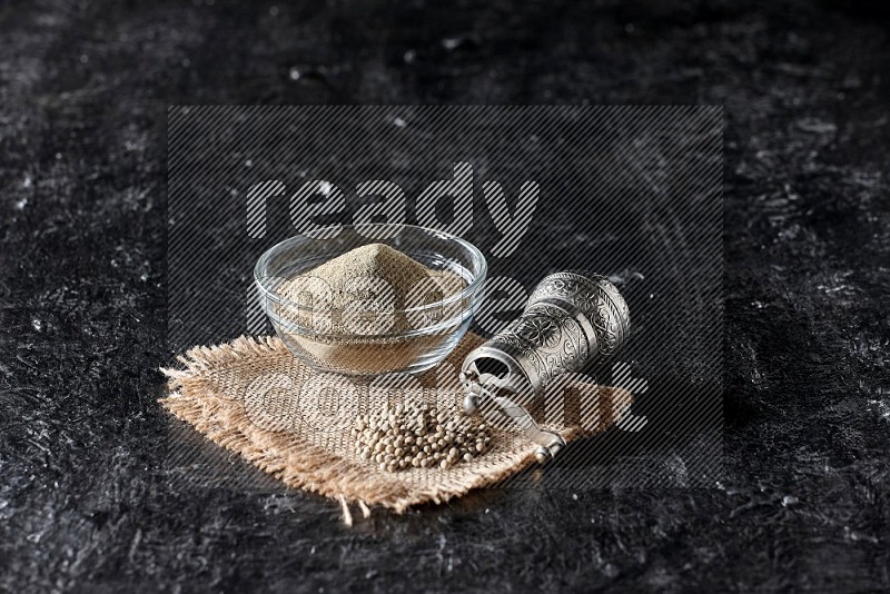 A glass bowl full of white pepper powder with white pepper beads on a burlap piece of fabric and a metal grinder on textured black flooring