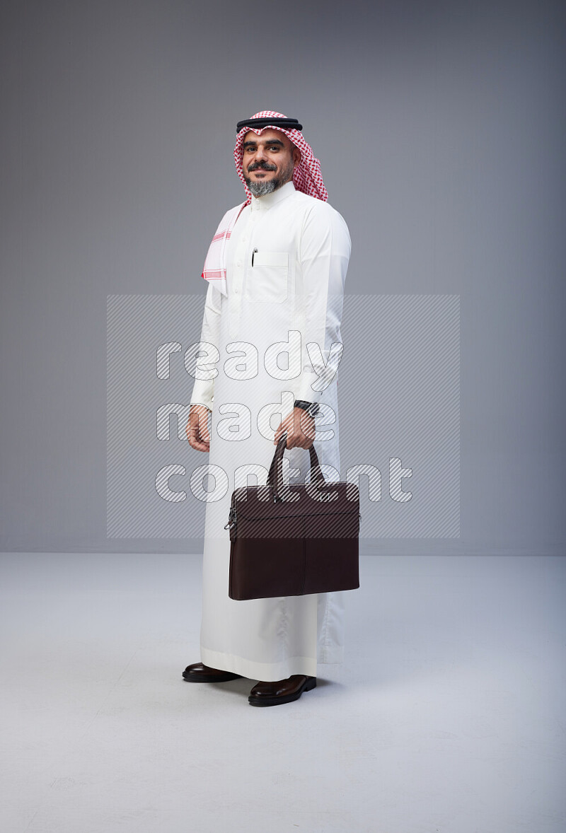 Saudi man Wearing Thob and red Shomag standing holding bag on Gray background