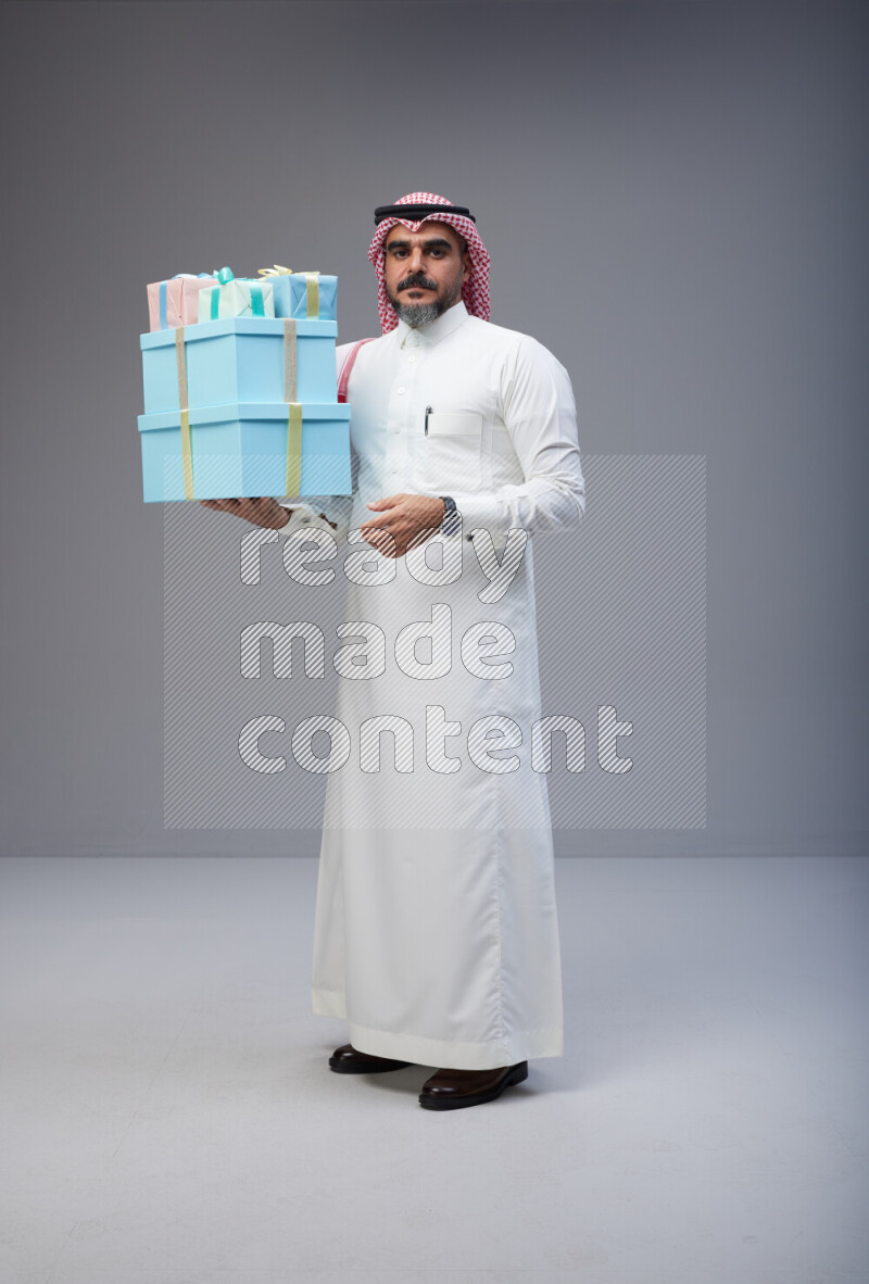 Saudi man Wearing Thob and red Shomag standing holding gift box on Gray background