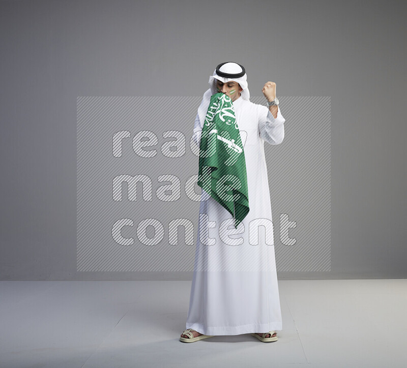 A Saudi man standing wearing thob and white shomag with face painting kissing big Saudi flag on gray background